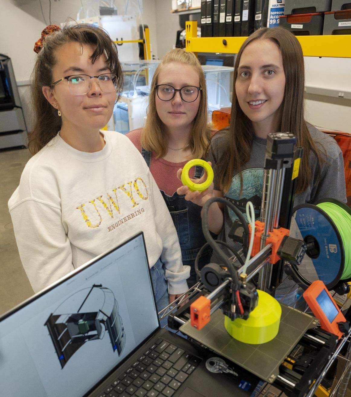 three senior design students stand behind a table displaying a digital model of their project, and a 3D printer that is creating a prototype.