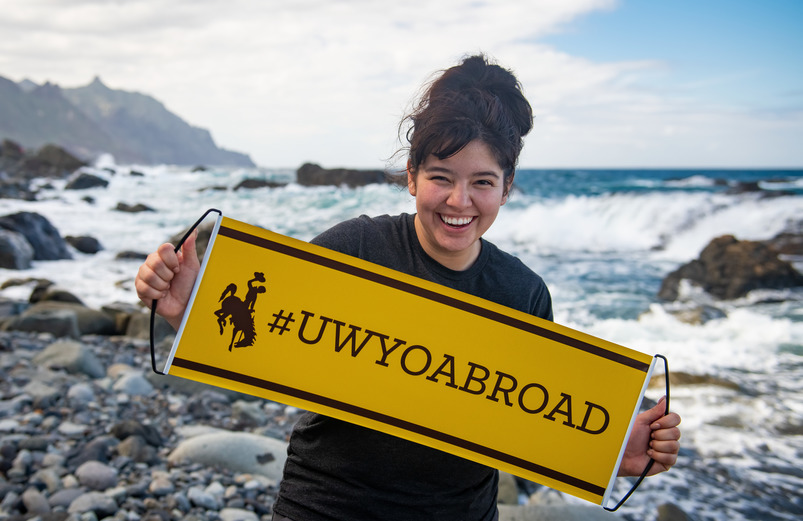 Girl holding UWYO Abroad sign in front of ocean