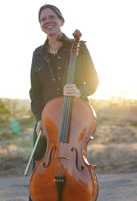Photo of Dr. Beth Vanderborgh outdoors, holding a cello in a jean jacket
