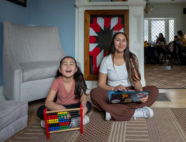 Female Native American student with daughter, holding laptop while daughter plays on an abicus