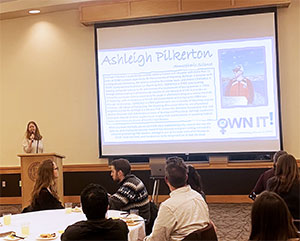 woman in front of seated group with large screen and projected image