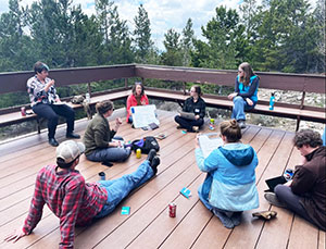 people sitting in a rough circle on a deck in a wooded area