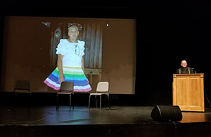 slide showing on a wall screen with man at podium reading