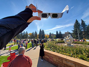 person holding eclipse viewing glasses with field of people in the background