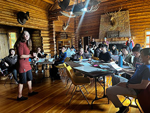 people at tables in wood-walled lodge with someone speaking