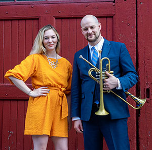 woman and man standing in front of a red door, with the man holding two trumpets