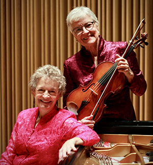 two women, one sitting and leaning on a grand piano and the other holding a viola
