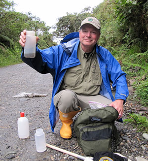 man crouching on a roadside, holding up a plastic bottle