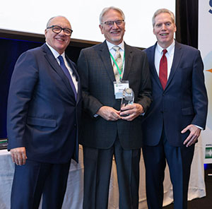 three men posing together, with the one in the middle holding an award