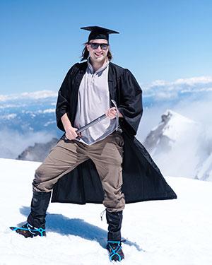 person standing in the snow on a mountain in a graduation cap and gown holding an ice-climbing pick