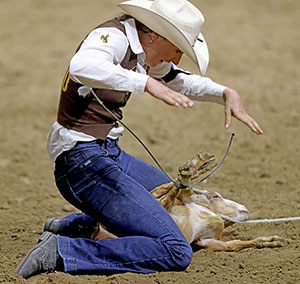 woman holding a goat with tied legs between her knees on the ground of an arena
