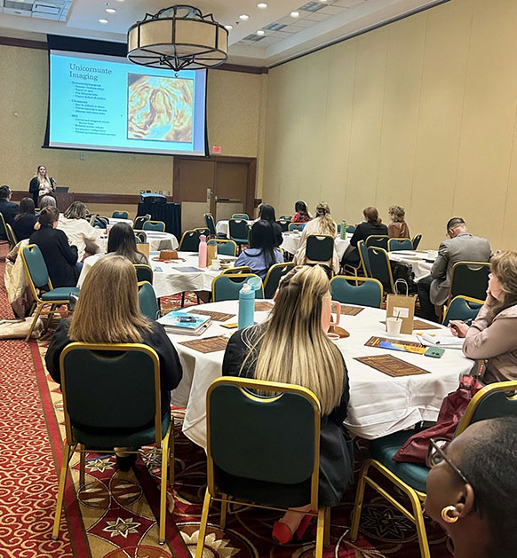 room full of people sitting at tables and looking at a screen high on a wall