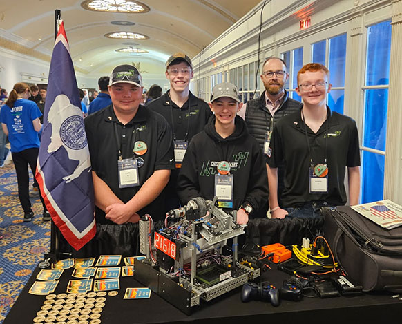 four teenagers and an adult standing behind a table with electronics on it