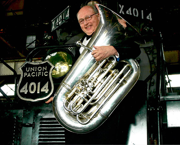 man holding a tuba while standing on the front of a steam engine