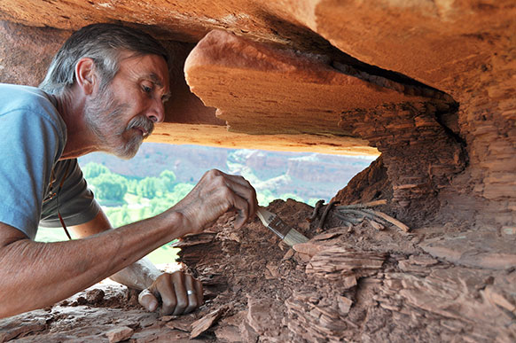 man crawling under a rock ledge using a paintbrush on the ground