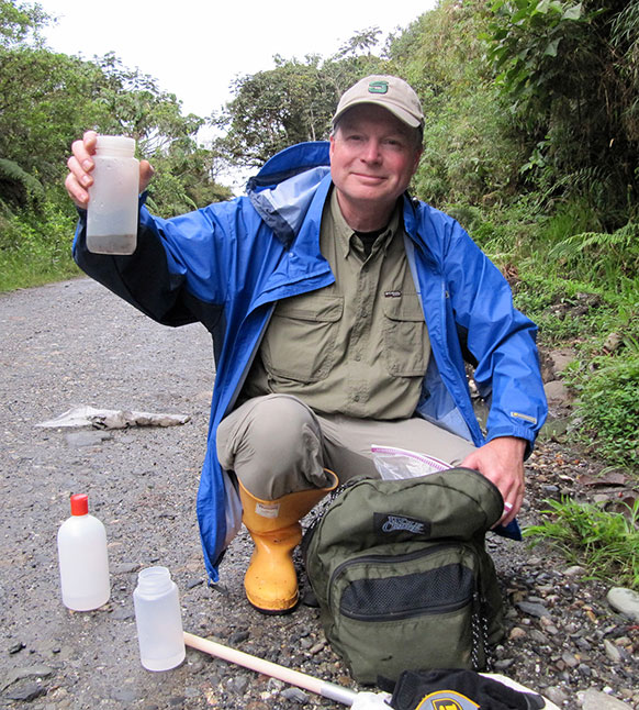 man squatting on a road in a forest holding up a container with water in it