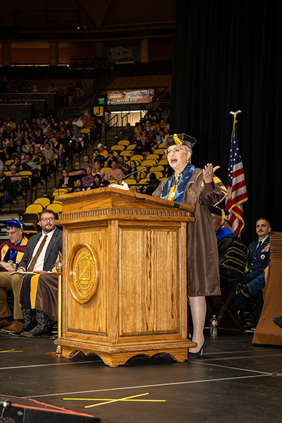 A woman stands behind a podium wearing a cap and gown