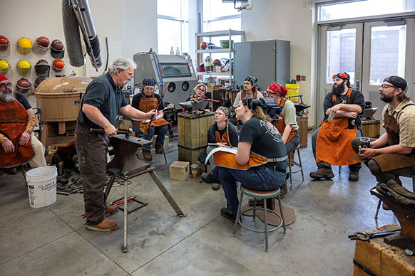 people in leather aprons watching a man at an anvil