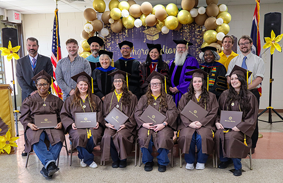 Women sit down wearing graduation gowns and hold their diplomas, with a group of people behind them.