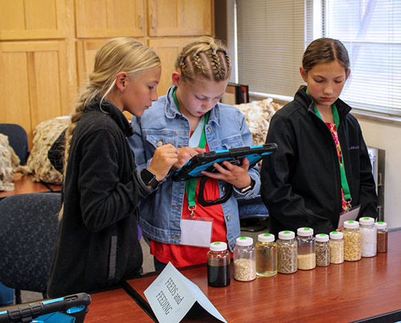 three girls holding a tablet while standing at a table holding small containers