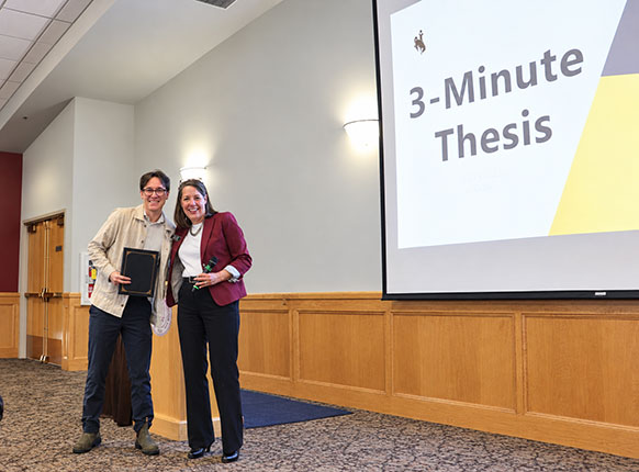 two people standing in a hall near three-minute thesis displayed on a screen
