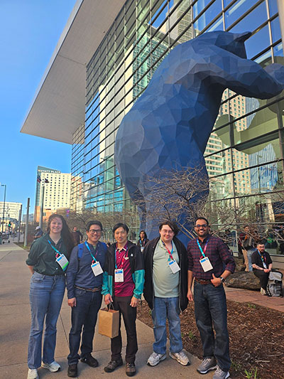 five people posing by a building with a huge blue bear sculpture looking into windows
