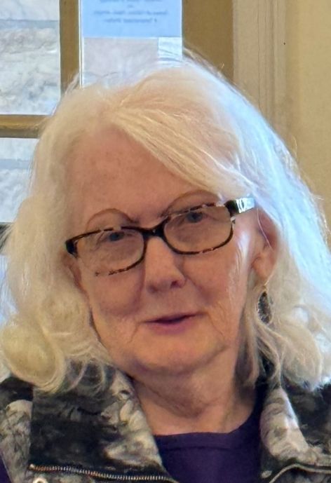 shirley boggs wearing glasses in a jacket and purple shirt standing in front of a window