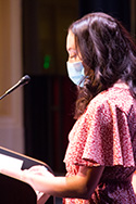 Masked young woman with dark long hair stands at microphone to speak