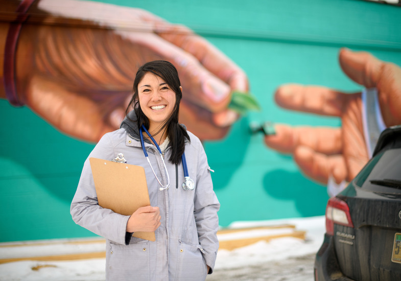 A graduate of a UW nursing program smiles in front of a local mural with a stethoscope and clipboard.