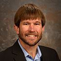 young man in suit with dark brown hair and fringe bangs, mustache and beard