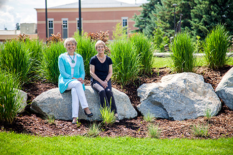 Two women sit on a boulder in lush green surroundings