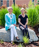 two women sit on a boulder amid luscious green backdrop