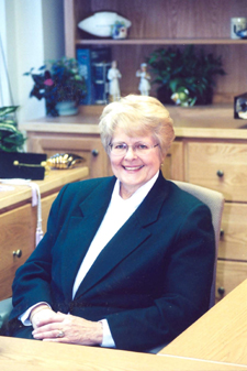 Professional woman with short blonde hair sitting at desk with memorabilia pictured behind