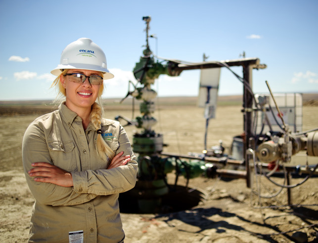 Woman in a hardhat and safety glasses standing outside facing the camera and smiling with oil extraction equipment in the background