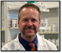 Man in white coat standing behind pharmacy counter with shelves of pill bottles 