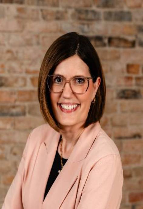 Smiling brown hair woman wearing glasses and a pink blazer with black shirt and gold necklace