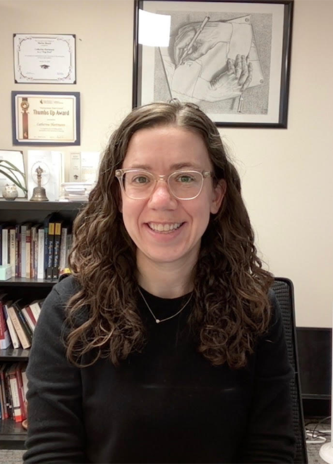 Smiling woman with wavy brown hair and glasses seated in an office.