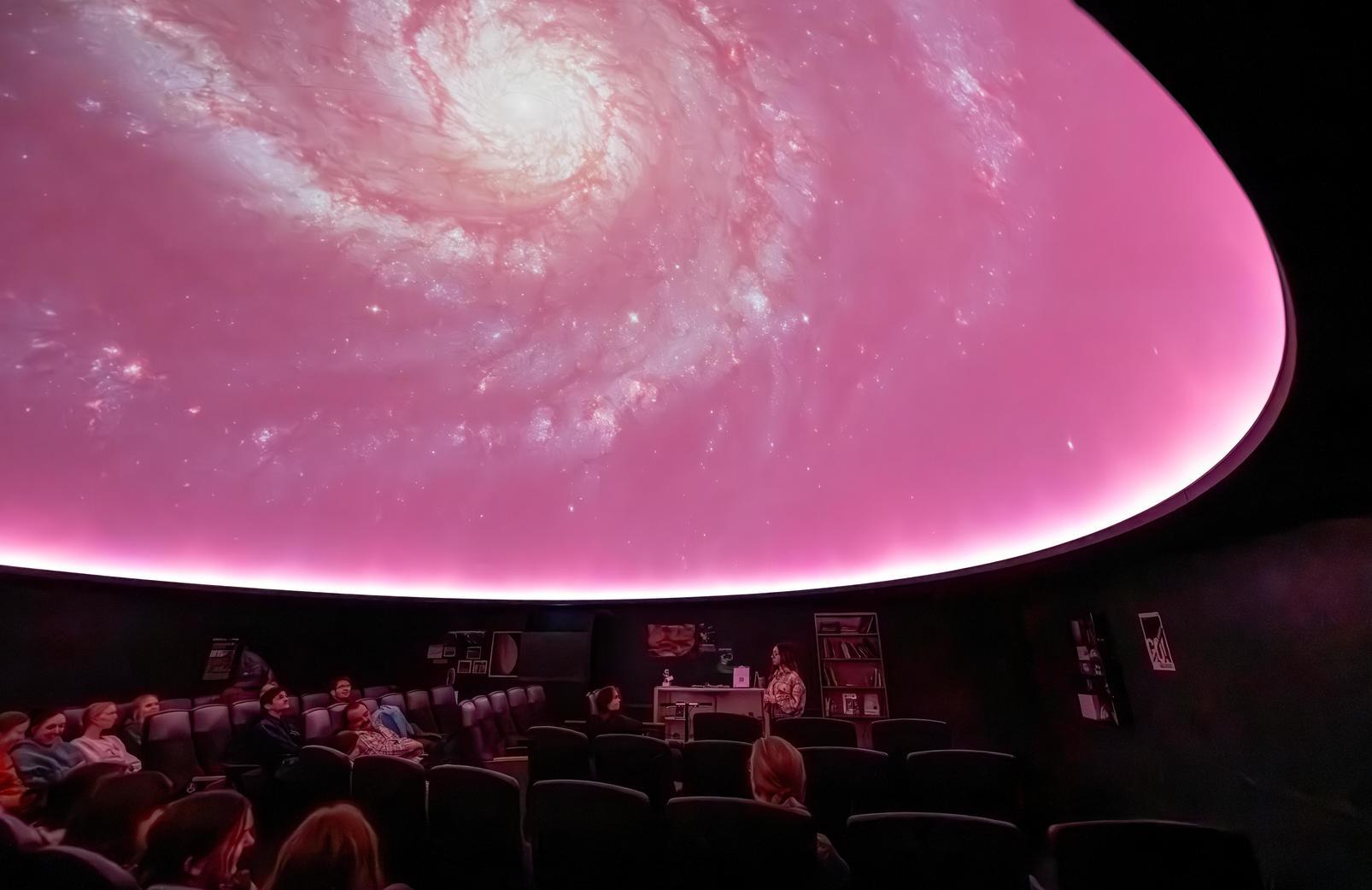 A group of people sit in the planetarium and look up at the ceiling while a show with two planets is displayed