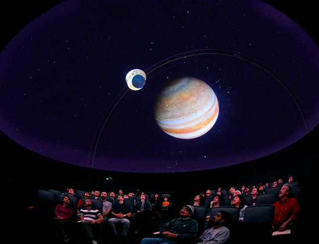 A group of people sit in the planetarium and look up at the ceiling while a show with two planets is displayed