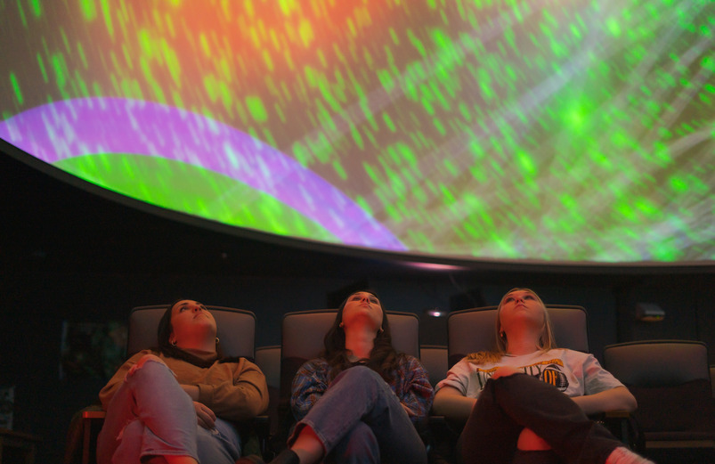 Three students sit in the UW planetarium and observe the images projected on the dome screen.
