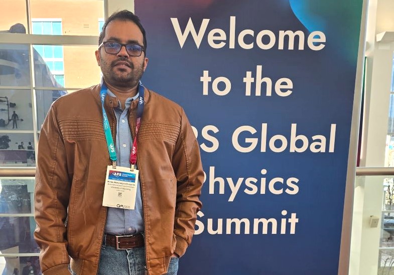 a young man with a conference badge stands in front of a standing banner for the Global Physics Summit