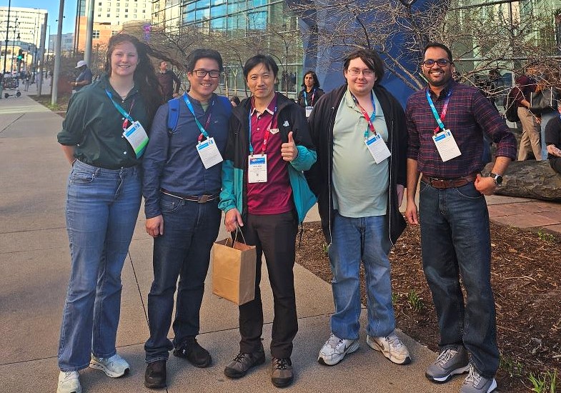 A group of five smiling people pose together outside in front of a building