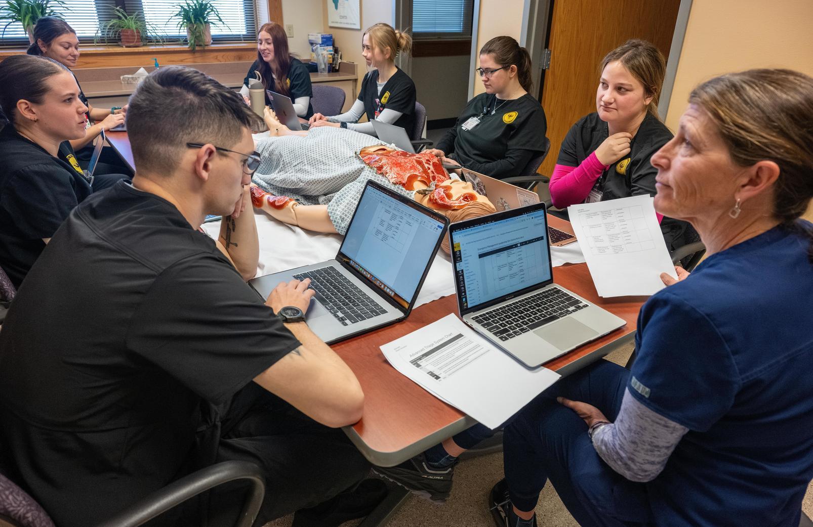 A group of nursing students sitting at a table with a simulated patient.