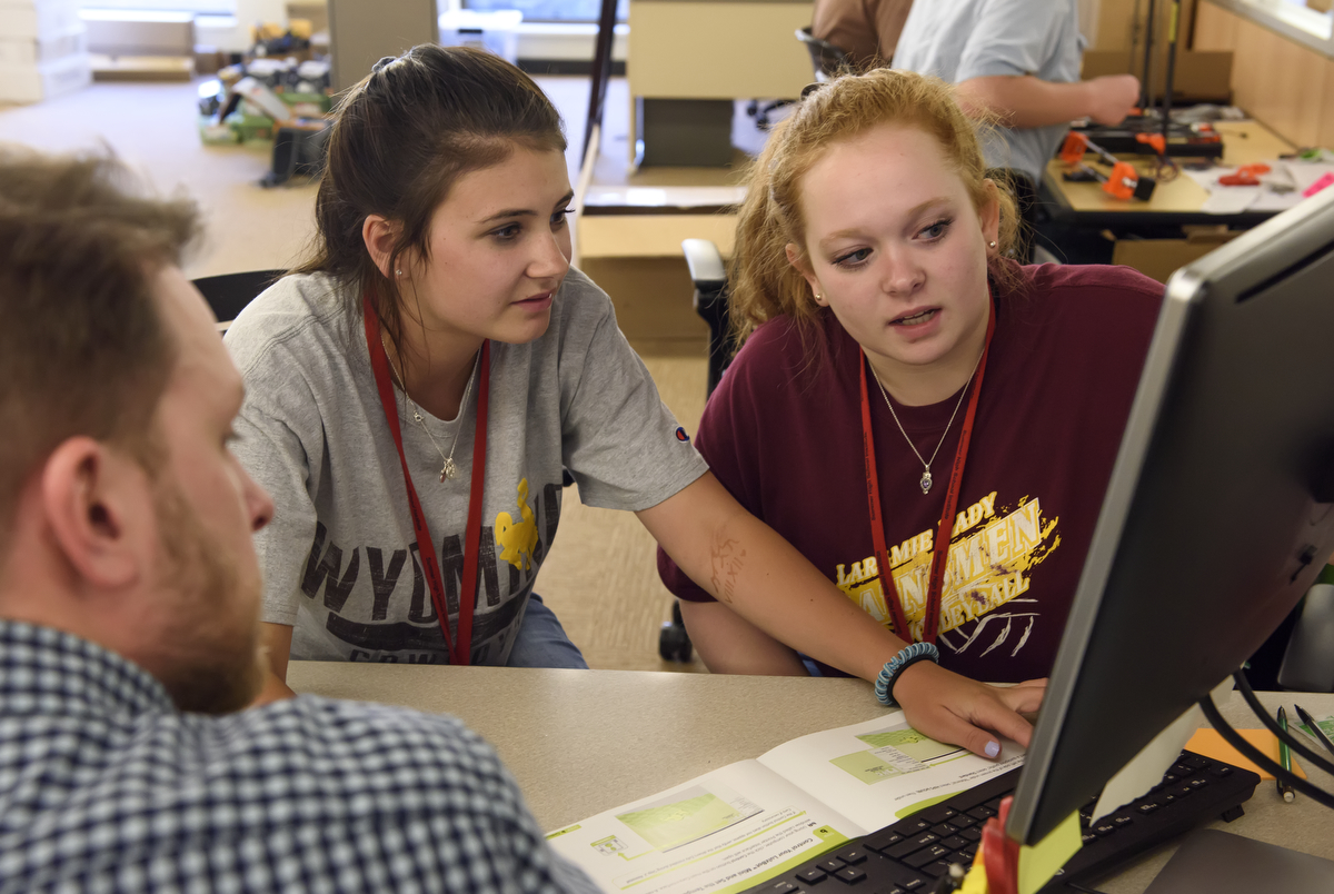 Three students looking at a computer while one of the three is pointing at a book