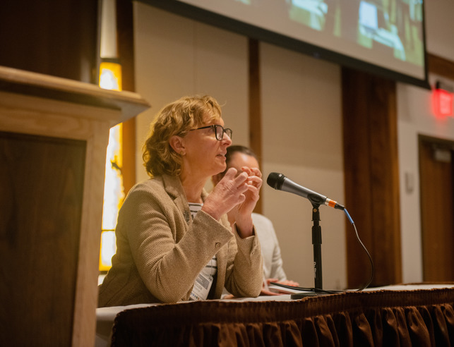 A speaker sits at a microphone podium