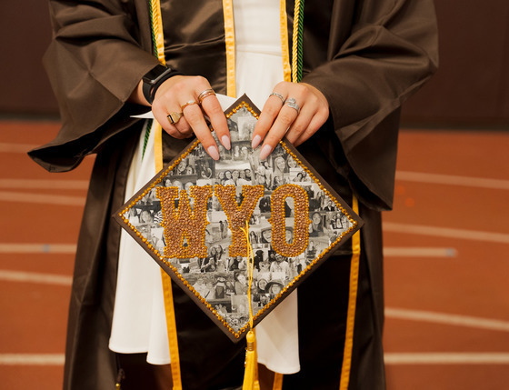 Student holding graduation cap decorated with images of friends and family and a large WYO printed in brown and gold in the center.