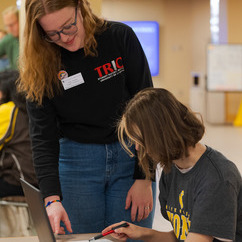 An advisor standing next to a student while they review a document on the table with a laptop open.