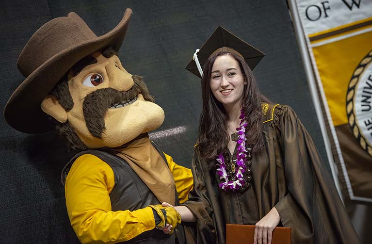 a graduating student holding a diploma and wearing graduation regalia with a purple orchid lei shakes hands with the UW mascot Pistol Pete