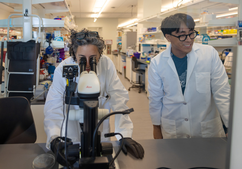 Two researchers in a lab, one smiling and one looking into a microscope