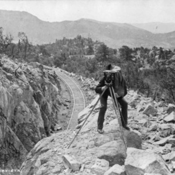 A black and white image of a man taking a landscape photo with mountains and a railroad behind him